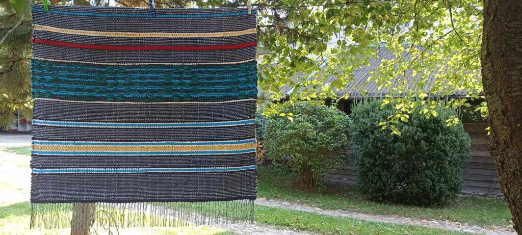 tkanina tzw. radziuszka z różnokolorowej włóczki zawieszona na sznurku w tle widać drzewa i krzewy.
