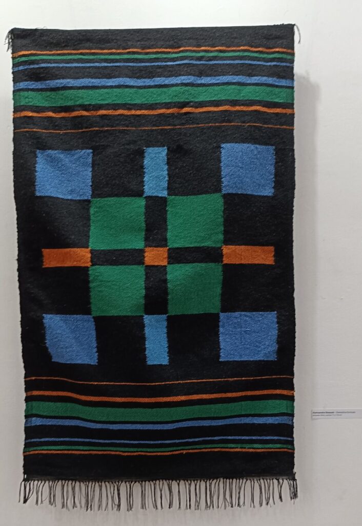 Tkanina wykonana metodą kilimy wisząca na ścianie. Kolory tkaniny czarna osnową i wątek w pasy czarno, pomarańczowo, niebiesko, zielone. Po środku wzór tzw. schematu tkackiego gwiazdki. Wykonała Aleksandra Staszak