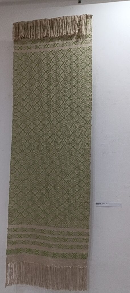 Tkanina wisząca na ścianie w kolorze białym. Osnowa w tkaninie szara bawełna, Zawątek bawełna metalizowana, wątek zielony len. Tkanina wykonana techniką przetykaną tzw. radziuszką z charakterystycznym motywem gwiazdy.